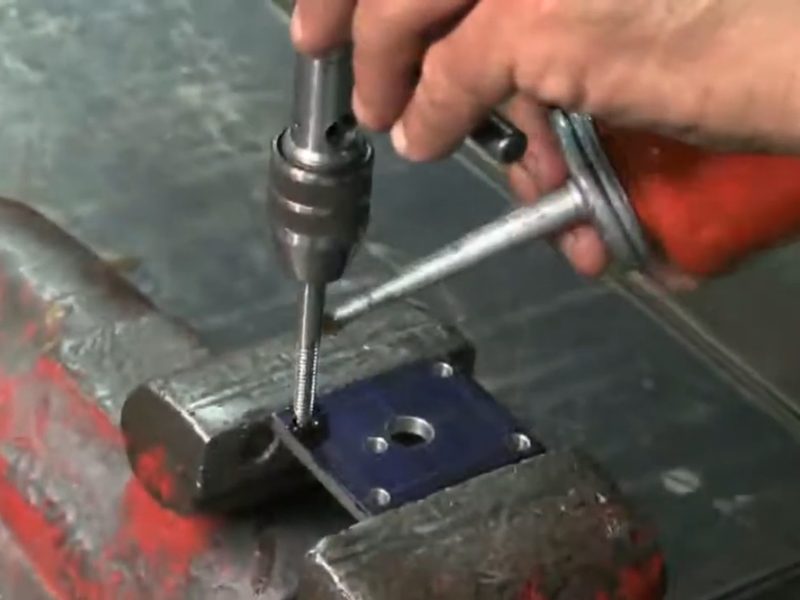 Hands starting a tap square into a layout-blued plate held in a
                      vise, using a T-handle tap wrench guided by a tapping block.