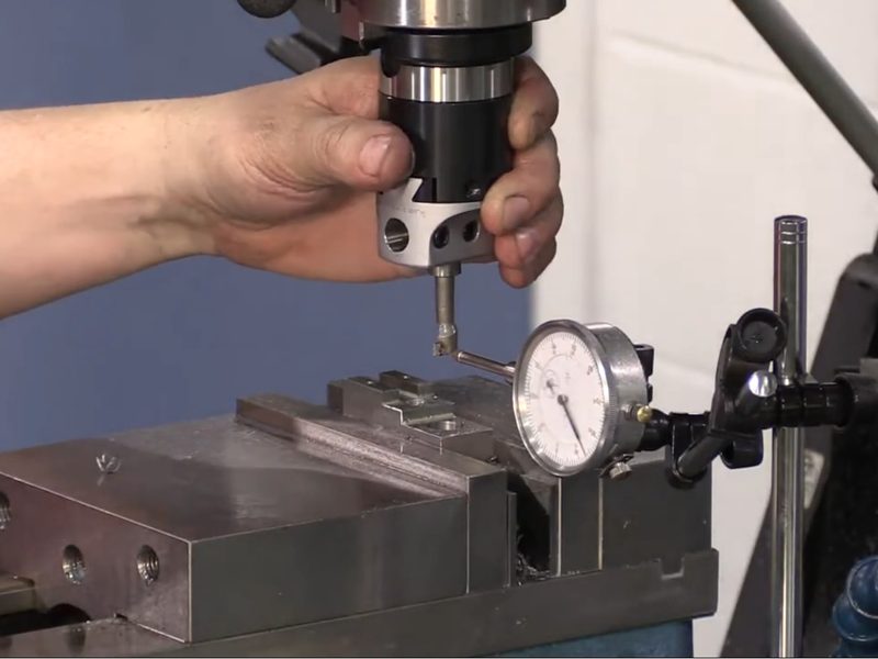 A hand turning the quill of a manual mill to bring an edge finder down
                      onto a workpiece clamped in a vise, with a dial indicator on a
                      magnetic stand reading against the vise jaw.