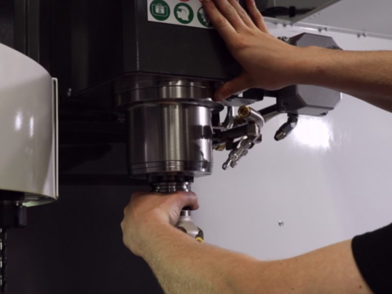 A student loading a tool holder into the spindle of a CNC machining
                      centre, one hand supporting the holder underneath while the other
                      presses the tool release button on the head.